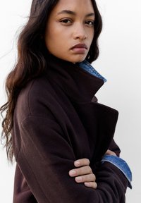 Ciemnobrązowa kurtka z wysokim kołnierzem, teksturowana tkanina, nałożona na jasnoniebieską koszulę dżinsową z widocznymi przeszyciami i mankietami.