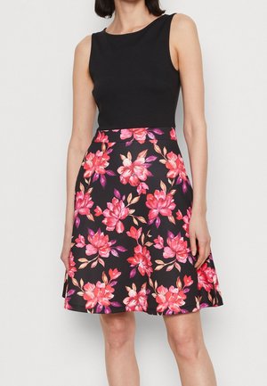 Femme portant une robe noire sans manches avec une jupe noire longueur genou présentant de grands motifs floraux rose vif et violet.