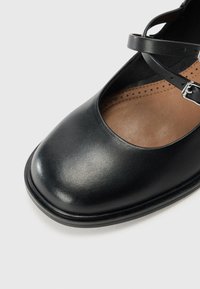 Black leather shoe with a rounded toe, cutout panels, and ankle strap featuring a silver buckle. Brown leather insole with perforations.
