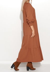 Robe longue, brune, à niveaux et manches bouffantes, en tissu doux, avec des accents froncés à la taille et à l'ourlet, associée à des bottes à talons brunes.