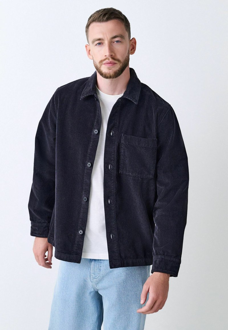 Navy Cordjacke mit Knopfleiste, Brusttasche und langen Ärmeln, kombiniert mit hellblauen Jeans. Glatte Textur, lässiges Design.