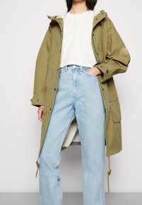 Długa kurtka w oliwkowej zieleni z regulowanym sznurkiem, w parze z białą, krótko przyciętą koszulą i jasnoniebieskimi szerokimi dżinsami; luźny krój, gładka tekstura.