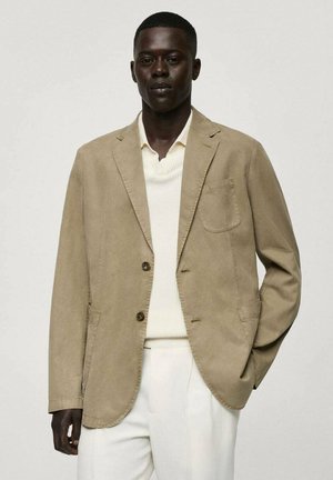 Blazer en coton beige avec une poche poitrine simple, deux boutons, revers crantés et une coupe légèrement décontractée, porté sur un pull crème et un pantalon blanc.