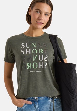 Jeune femme portant un t-shirt graphique vert olive "Sunny Shore" et tenant un sac fourre-tout noir sur son épaule.