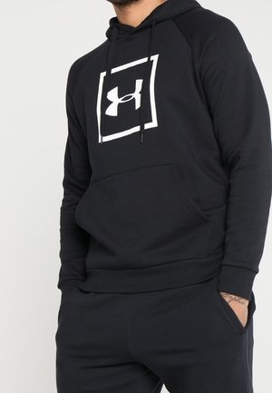 Homme portant un sweat à capuche noir Under Armour et un pantalon noir les mains dans les poches, montré de la poitrine aux hanches sur fond blanc.