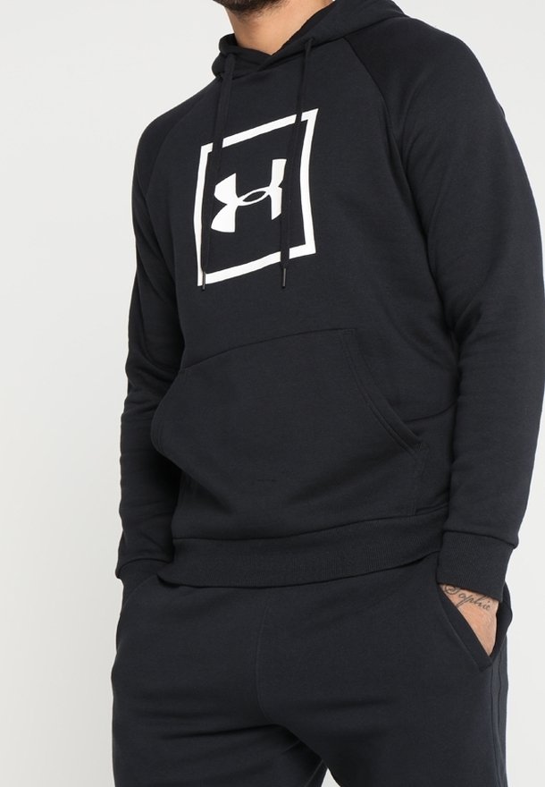 Homme portant un sweat à capuche noir Under Armour et un pantalon noir les mains dans les poches, montré de la poitrine aux hanches sur fond blanc.
