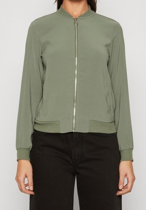 Femme portant une veste bomber vert olive clair zippée, avec poignets et ourlet côtelés, associée à un pantalon noir.