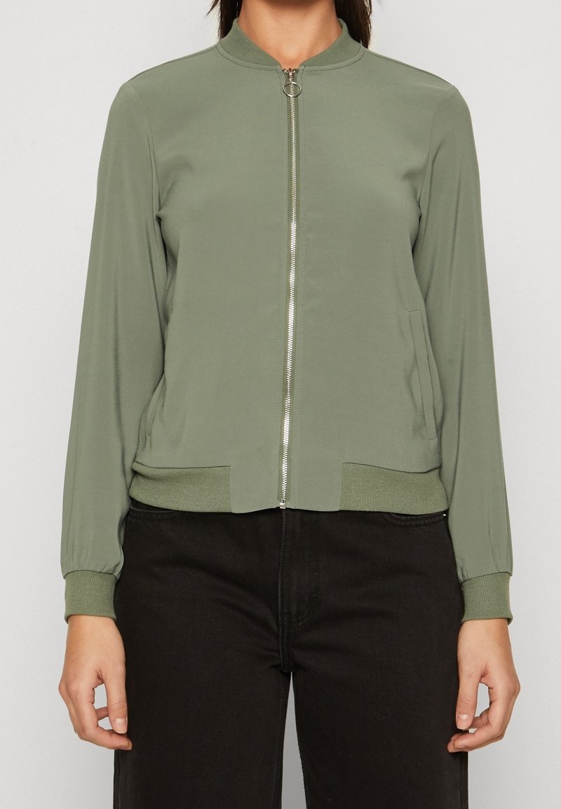 Femme portant une veste bomber vert olive clair zippée, avec poignets et ourlet côtelés, associée à un pantalon noir.