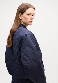 Marineblå quilted bomberjakke med et strukturert mønster, ribbestrikket krage og glidelås. Har en avslappet passform og polstret design.