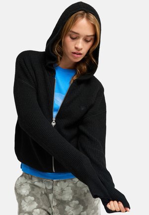 Junge Frau trägt einen schwarzen Kapuzen-Reißverschluss-Pullover über einem blauen Hemd und grauen Hosen mit Blumenmuster, schaut nach unten und richtet ihren Ärmel.
