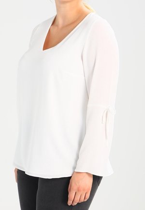 Femme portant un chemisier blanc à manches longues avec un col en V et un détail noué sur la manche, associé à un pantalon noir.