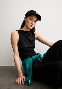 Top senza maniche nero con una consistenza liscia, abbinato a pantaloni neri larghi. Un maglione a coste teal è annodato in vita. Cappellino nero visibile.