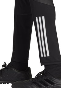 Zwarte sportbroek met een tapered ontwerp, meshdetails en drie witte strepen aan de zijkant. Gecombineerd met zwarte sportschoenen.
