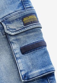 Close-up van blauw denimstof met een zak en een geel label met de tekst "Next Supply Authentic Durable Goods."