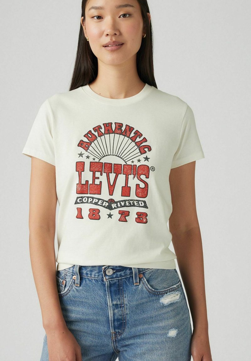 T-shirt en coton de couleur crème avec un graphique rétro du logo Levi's rouge, accompagné d'étoiles et de texte. Associé à un jean en denim bleu usé.