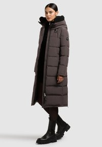 Cappotto imbottito lungo di un grigio scuro con colletto alto, chiusura con zip e stivali neri robusti. Presenta un design trapuntato e una fodera interna strutturata.