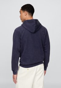 Tengerészkék pulóver puha anyagból, állítható zsinóros kapucnival és raglán ujjakkal, világos színű nadrággal párosítva.