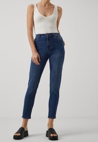 Blå högmidjade jeans med smal passform, gjorda av denim, kombinerade med en vit ribbad linne och svarta tjocka slide-sandaler.