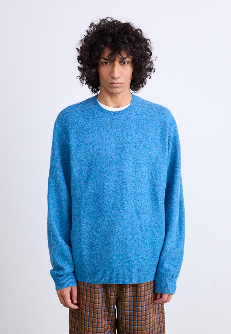 Pull bleu en tissu texturé doux, avec un col rond et des poignets côtelés. Un petit logo est visible sur la poitrine. Pantalons à carreaux bruns.