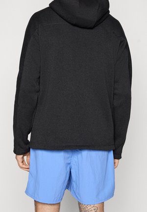 Osoba nosząca czarną bluzę z kapturem i jasnoniebieskie spodenki, pokazana od tyłu na jednolitym tle.