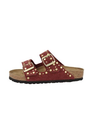 Sandale à glisser en suède rouge avec deux sangles ajustables, des accents en studs dorés et un footbed en liège texturé. Semelle extérieure en caoutchouc noire.
