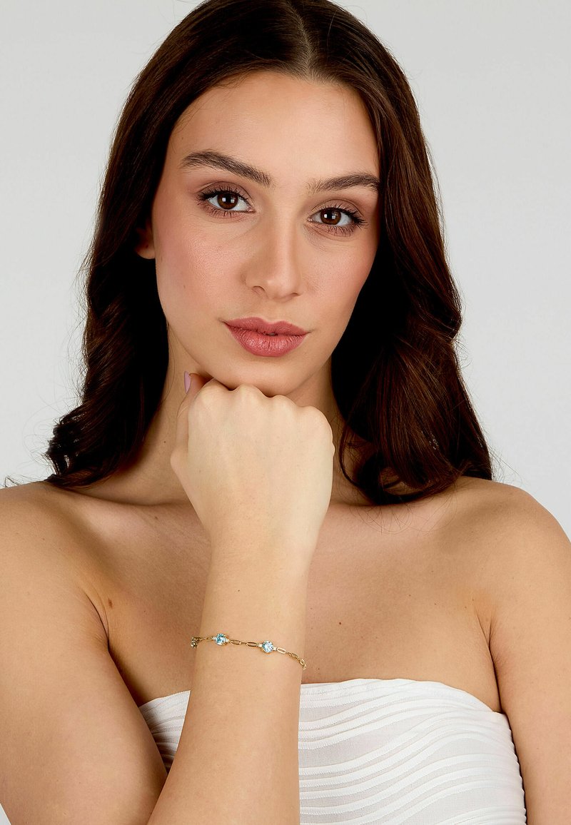 Bracciale d'oro con due pietre azzurre chiare, collegate da una delicata catena. Modella che lo indossa al polso, braccia incrociate su uno sfondo semplice.
