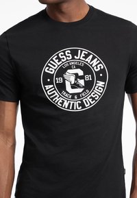 Sort bomulds t-shirt med et hvidt rundt logo, der har "Guess Jeans," "Los Angeles," og en grafik af en vingeret fod, 1981.