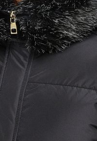 Detail černé zimní bundy s lesklým umělým kožíškem a viditelným zlatým jezdcem na zipu.