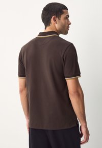 Brun poloshirt med en tekstureret finish, med krave og korte ærmer. Fremhævet med gule og hvide striber på ærmekanterne.