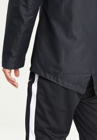 Personne portant une veste noire et un pantalon de sport noir avec une bande blanche sur le côté, debout, le bras gauche détendu le long du corps.