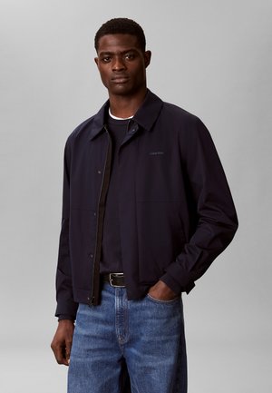 Mand iført navyblå lynlåstrøje, sort skjorte, blå jeans og sort bælte, stående mod en ensfarvet grå baggrund.
