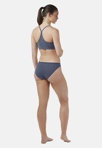 Mujer de pie descalza, de espaldas, con un sujetador deportivo azul de tirantes finos y ropa interior azul a juego, contra un fondo plano.
