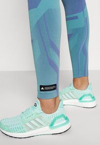 Mintgröna löparskor med meshuppdel, vit Boost-sula och ljusblå detaljer, tillsammans med åtsittande leggings med geometriska mönster i nyanser av blått.