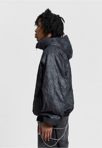 Schwarze, zerknitterte Stoffjacke mit Kapuze, ausgestattet mit einem Reißverschluss und elastischen Bündchen. Kombiniert mit dunklen Jeans und einem silbernen Kettenakzent.