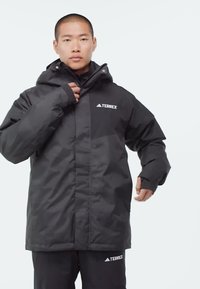 Muž zapínající černou zimní bundu adidas Terrex, v odpovídajících černých kalhotách Terrex, na jednoduchém bílém pozadí.