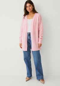Cardigan en maille rose clair avec col en V, coupe oversize et poignets côtelés, associé à un jean bleu taille haute et des ballerines beiges à bout pointu.