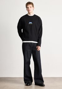 Μαύρη μπλούζα με μπλε και κίτρινο λογότυπο στο στήθος, συνδυασμένη με φαρδιά σκούρα τζιν. Το μοντέλο στέκεται μπροστά σε ουδέτερο φόντο.