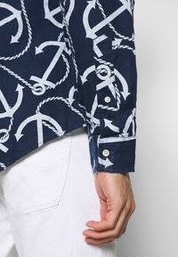 Chemise bleu marine avec un motif ancre et corde blanche, manches longues avec poignets à bouton, fabriquée en tissu texturé, associée à un pantalon blanc.