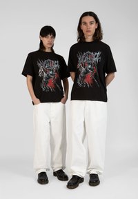 Tricouri grafice negre cu un design roșu și argintiu, asortate cu pantaloni largi albi și pantofi negri. Fundal minimalist.