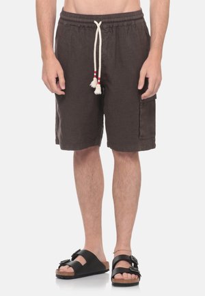 Uomo che indossa pantaloncini neri con laccetto bianco e sandali neri, in piedi su uno sfondo bianco uniforme. È visibile solo la parte inferiore del corpo.