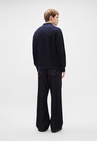 Pulôver azul-marinho tricotado com um acabamento texturizado, combinado com jeans escuros de perna larga. O conjunto apresenta um corte descontraído e punhos canelados.