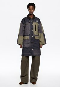Lange, oversized jas gemaakt van een mix van donkergrijze en olijfgroene stof, met grote zakken, een corduroy kraag en drukknoopsluitingen.