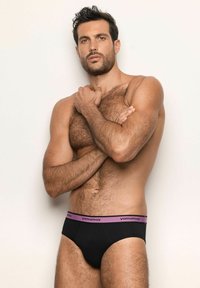 Muskulöser Mann mit dunklen Haaren posiert oben ohne, trägt schwarze Unterwäsche mit einem lila Bund, auf dem "yamamay" steht, vor einem schlichten, hellen Hintergrund.