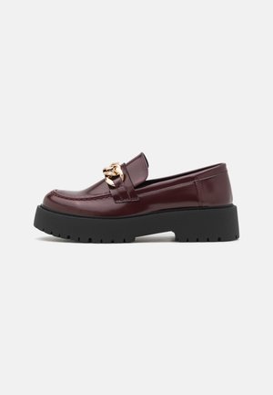 Mocassin en cuir bordeaux avec semelle noire épaisse et détail en chaîne dorée sur le dessus, présenté de côté sur un fond blanc.