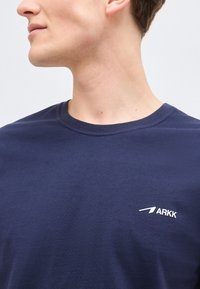 Νέος άνδρας που φοράει μπλε σκούρο κοντομάνικο μπλουζάκι ARKK, κοιτάζοντας προς τα αριστερά, με εμφανή τον λαιμό, το πηγούνι και το πάνω μέρος του στήθους.