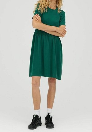 Femme portant une robe verte à manches courtes jusqu'aux genoux, des chaussettes blanches et des chaussures de sport noires, debout les bras croisés.