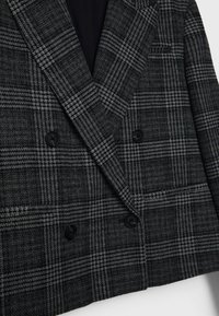 Blazer gris foncé croisé avec motif à carreaux noir et blanc et boutons noirs, présenté sur fond blanc.