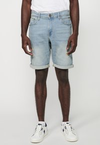 Shorts en denim bleu clair avec un ourlet retroussé, présentant des détails déchirés et un design à cinq poches, associés à des baskets blanches.