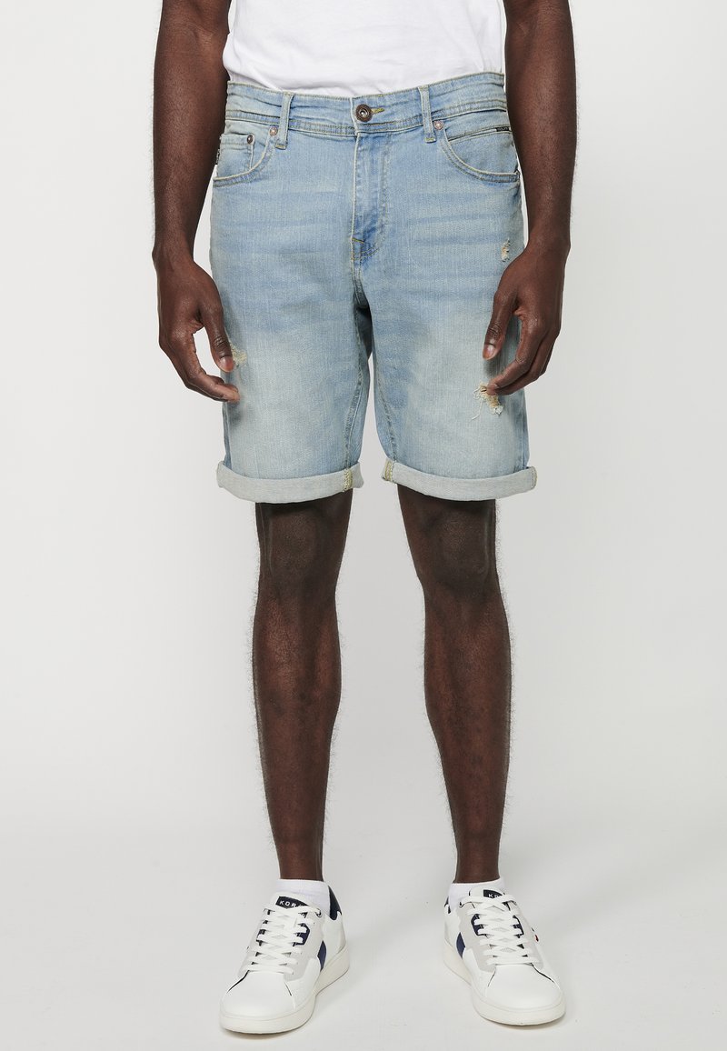 Shorts en denim bleu clair avec un ourlet retroussé, présentant des détails déchirés et un design à cinq poches, associés à des baskets blanches.