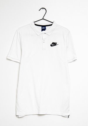 Biała koszulka polo Nike z krótkim rękawem, z czarnym logo po lewej stronie na piersi, zawieszona na czarnym wieszaku na białym tle.